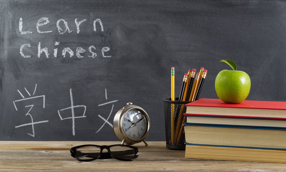 Student Learning Chinese With Books, Pencils, Clock, Reading Glasses And An Apple In Front Of Chalkboard With Mandarin Text.
