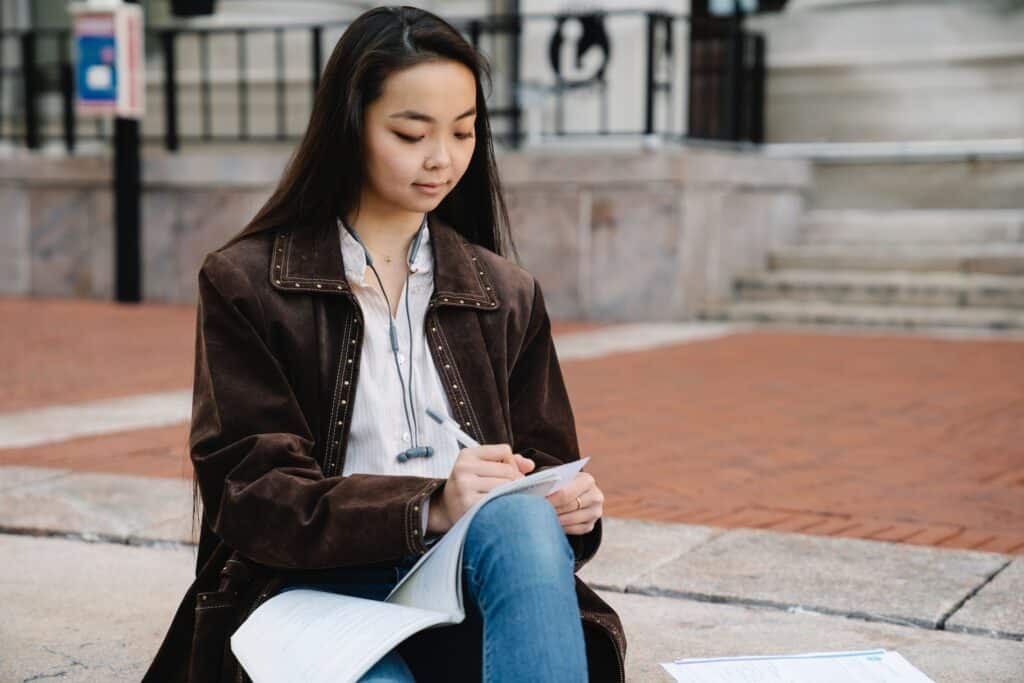 woman-of-asian-descent-studying-while-sitting-on-concrete-floor-outside-of-building