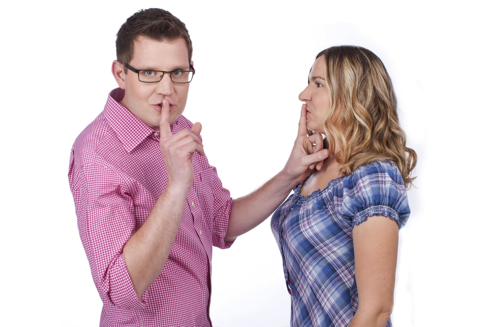 man-in-pink-polo-shirt-putting-one-finger-on-his-lips-and-another-finger-on-woman-in-blues-lips-to-silence-them-both