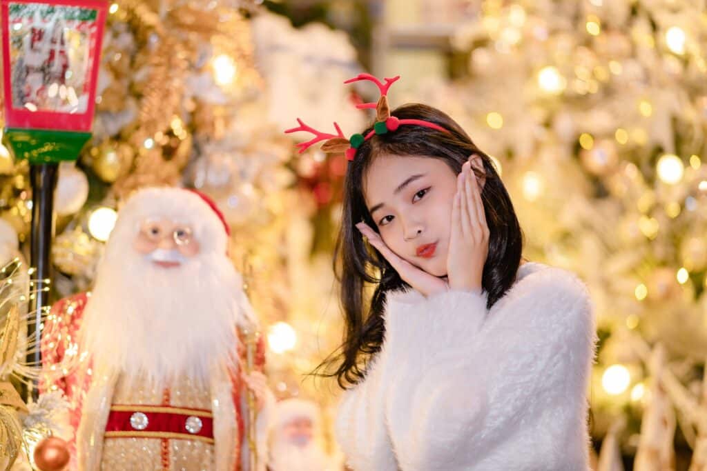 asian-woman-in-white-fluffy-jacket-and-fake-red-antlers-cupping-face-in-hands-and-making-cute-pose-against-christmas-background
