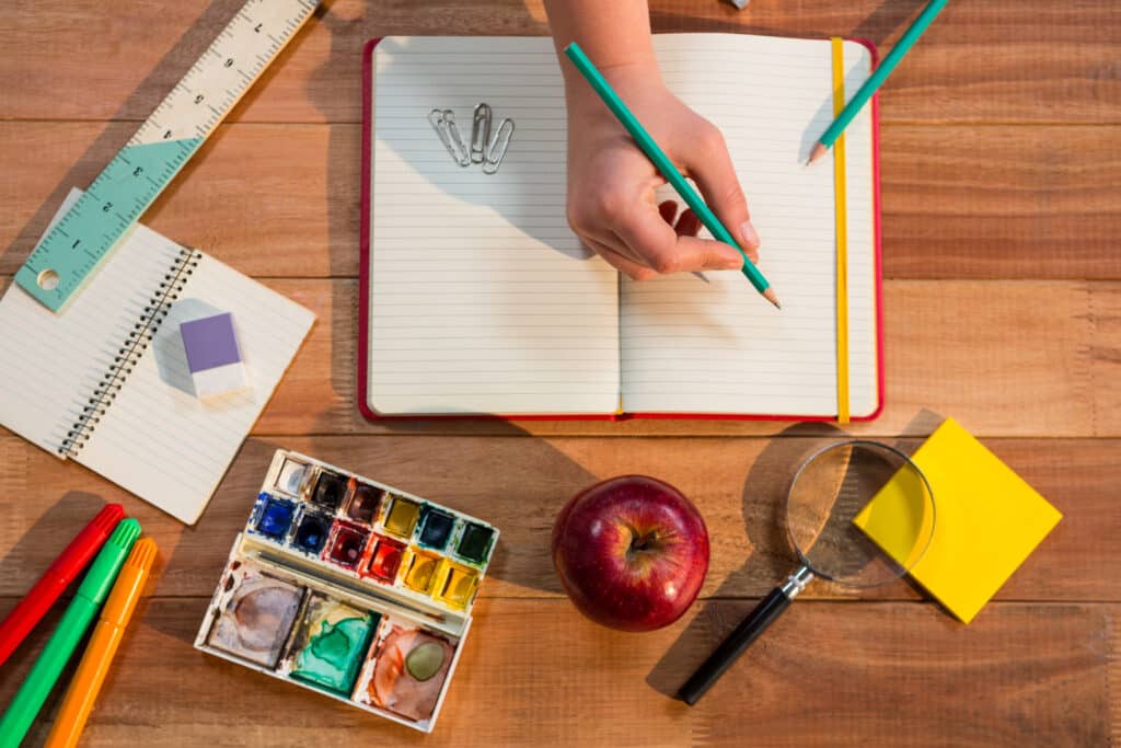 overhead-shot-of-hand-holding-pencil-over-blank-notebook-surrounded-by-various-school-supplies