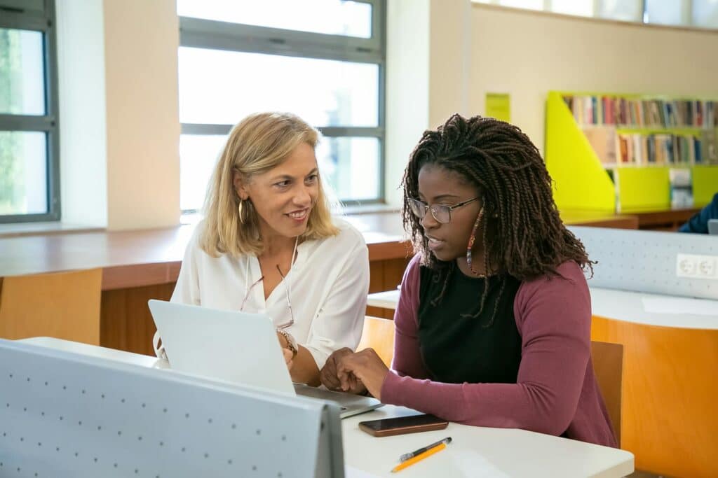 blond-teacher-discussing-something-on-white-laptop-with-student-of-african-descent