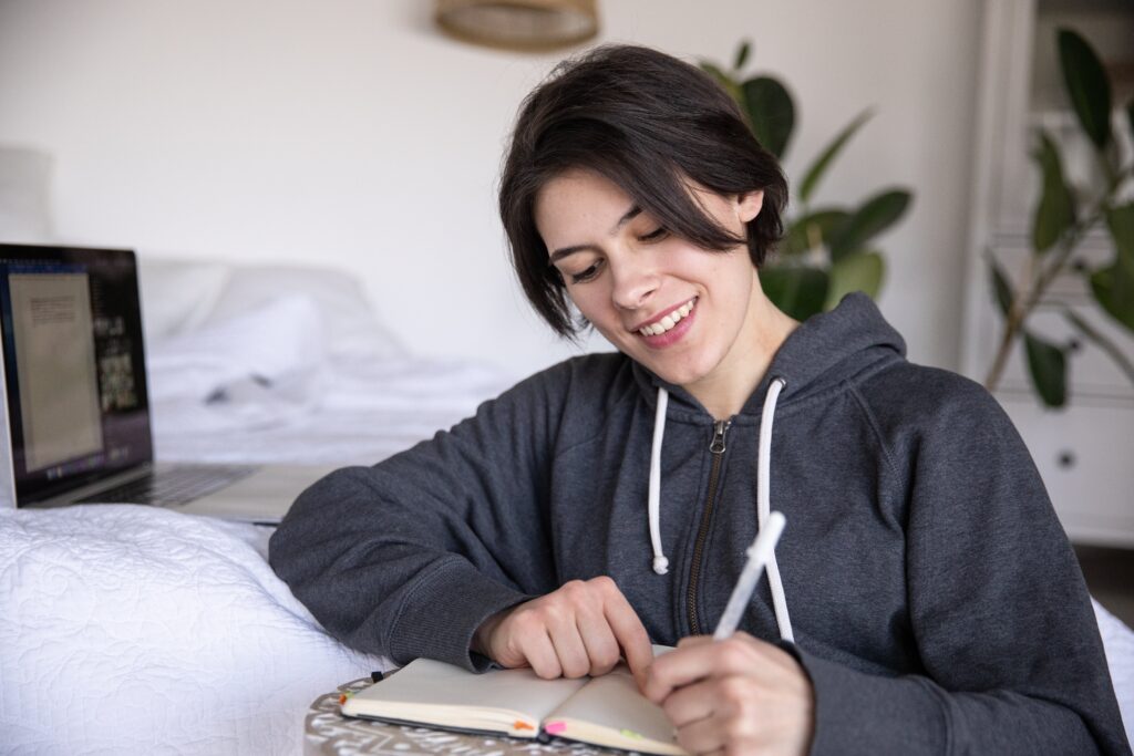 woman-in-gray-hoodie-smiling-as-she-writes-in-her-notebook