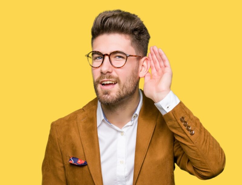 young handsome business man wearing glasses smiling with hand over ear listening against a yellow background