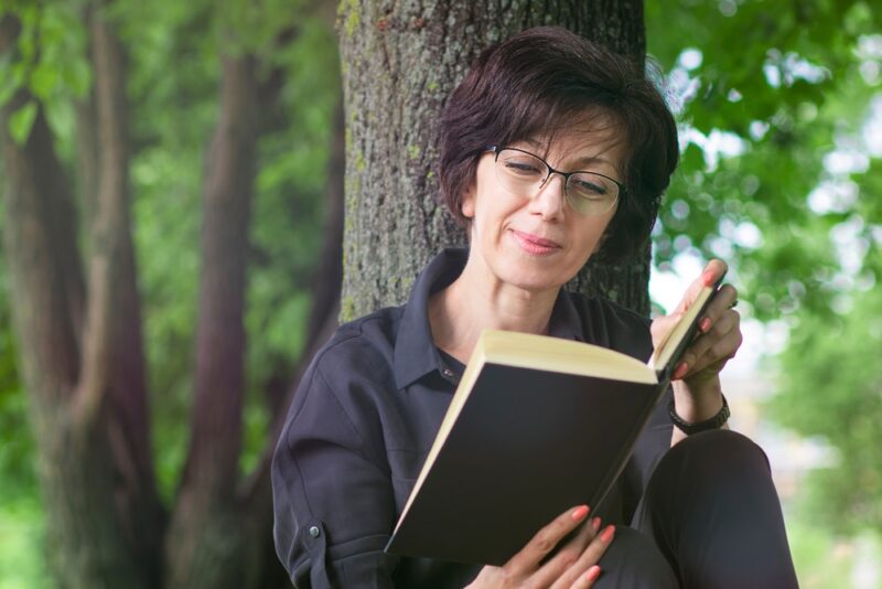 Beautiful elderly mature adult woman sitting on the ground, grass in the summer park, reading an interesting book in glasses, leaning on a tree, smiling