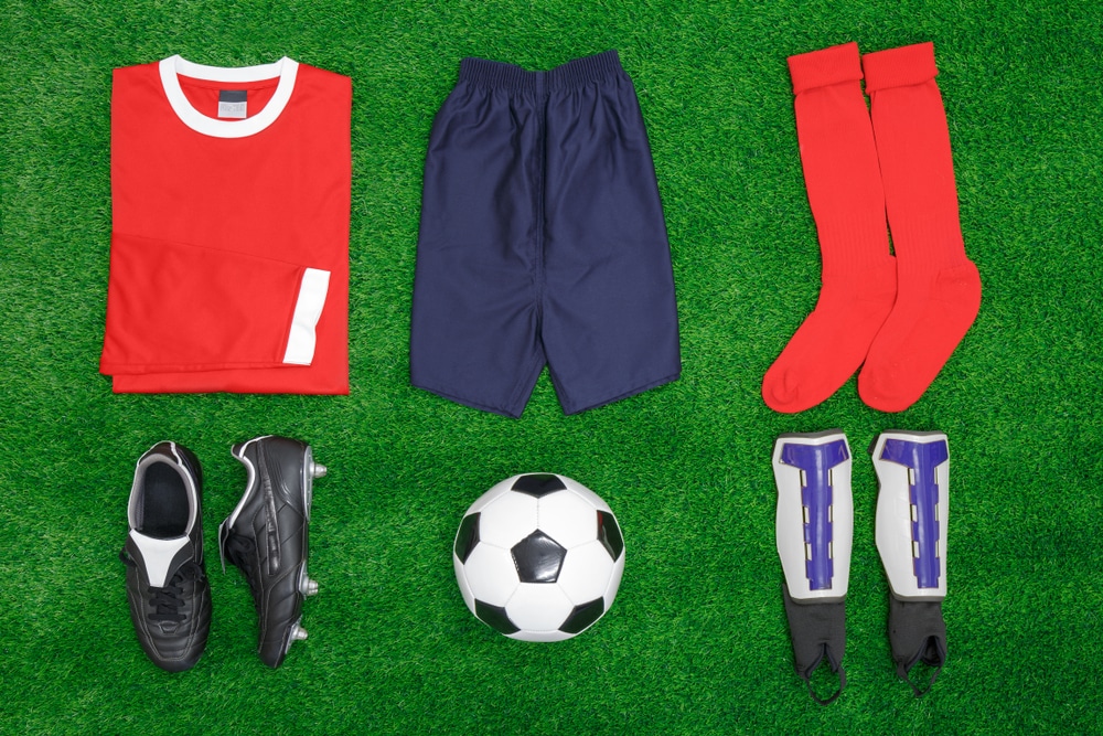 A flat lay arrangement of football or soccer kit on grass, with shirt, shorts,socks, boots, shin pads and ball.
