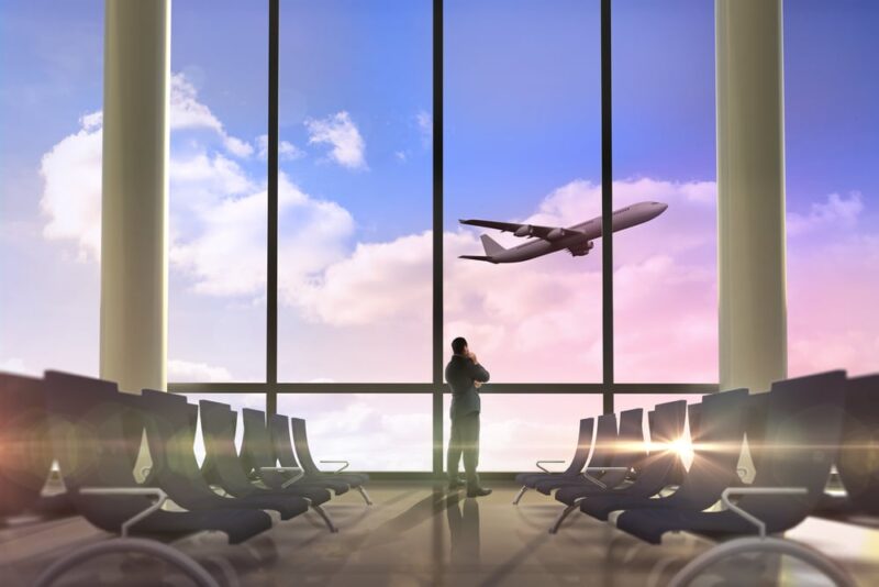 man looking out a window at an airport watching an airplane take off