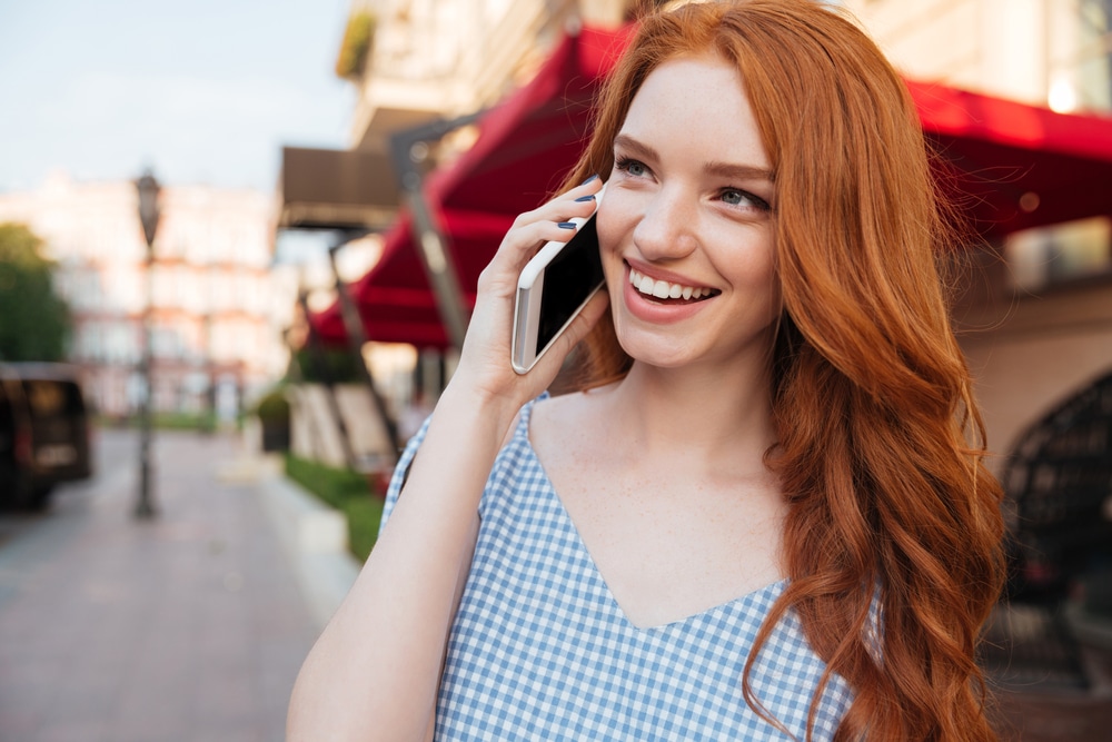 close up photo of smiling girl with long red hair talking on mobile phone while standing outdoors on a city street