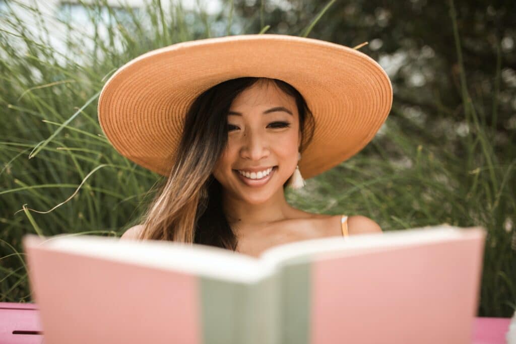 smiling-woman-of-asian-descent-wearing-wide-brimmed-hat-reading-pink-book