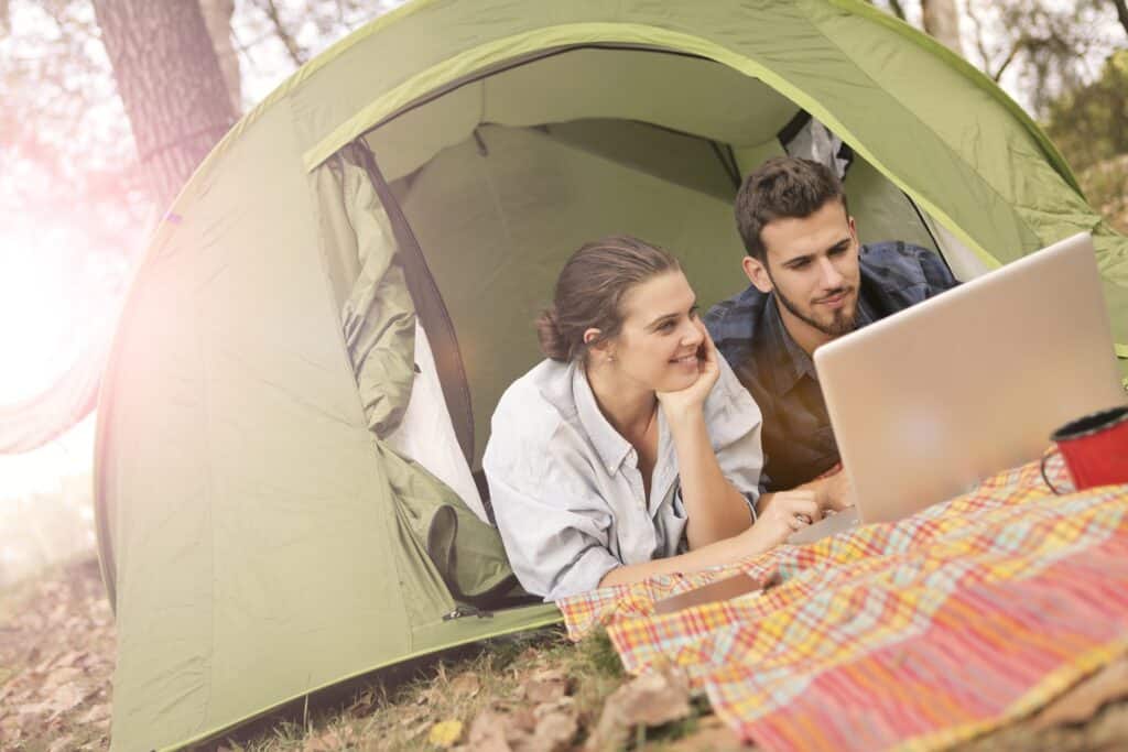 diagonal-shot-of-happy-couple-using-white-laptop-in-green-tent-with-sun-shining-in-background