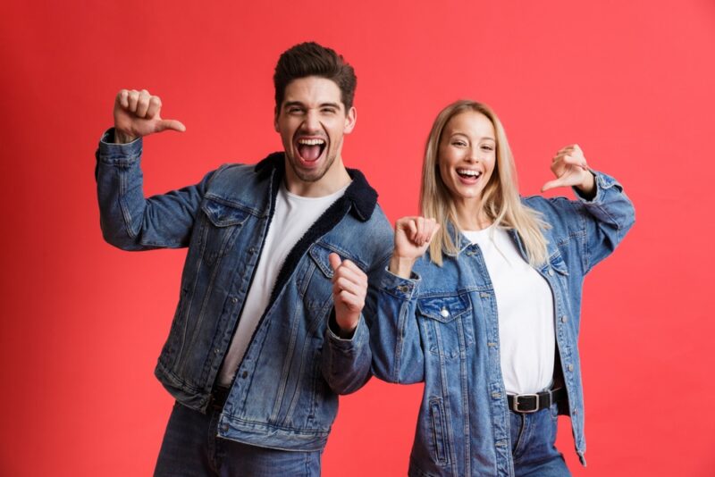 Portrait of a happy young couple dressed in denim jackets standing together isolated over red background, pointing at themselves