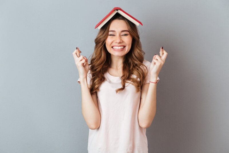a happy girl holding a book on her head with crossed fingers over gray wall background