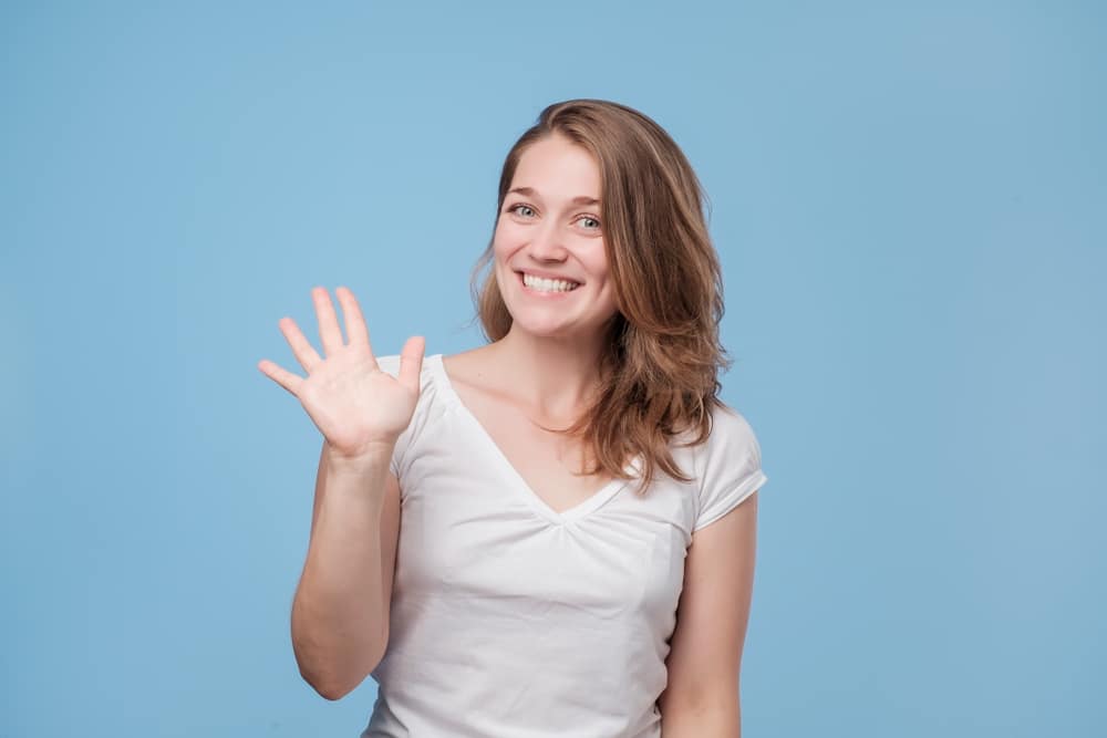 Attractive friendly looking young woman smiling happily, saying Hello, Hi or Bye, waving hand. Showing five fingers. Studio shot