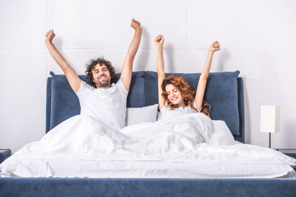 husband-and-wife-waking-up-in-bed-smiling-with-arms-outstretched-in-v-shape-towards-ceiling