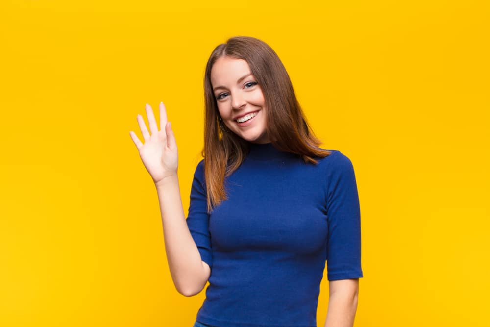 young red head woman smiling happily and cheerfully, waving hand, welcoming and greeting you, or saying goodbye against flat wall