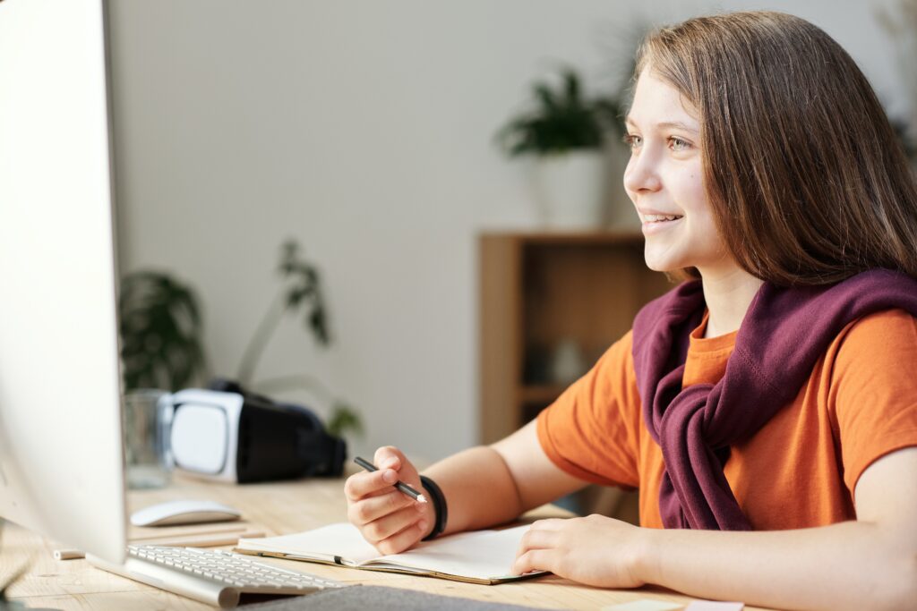 photo-of-a-girl-studying-and-smiling-at-a-computer-while-taking-notes