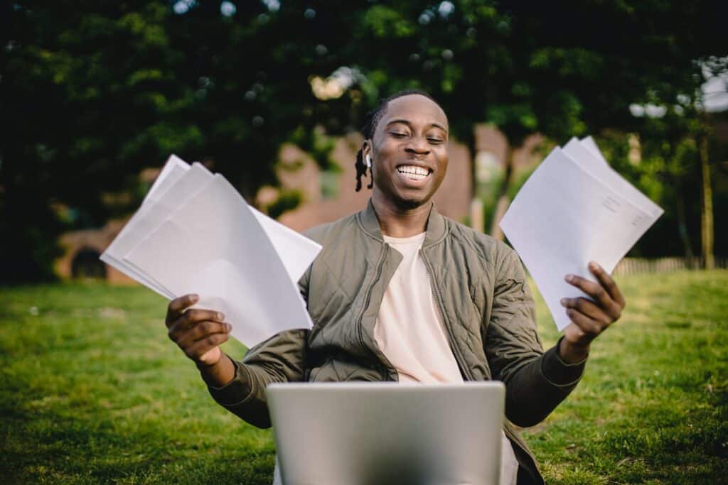 smiling-man-of-african-descent-holding-papers-in-both-hands-as-he-looks-at-laptop-in-front-of-him