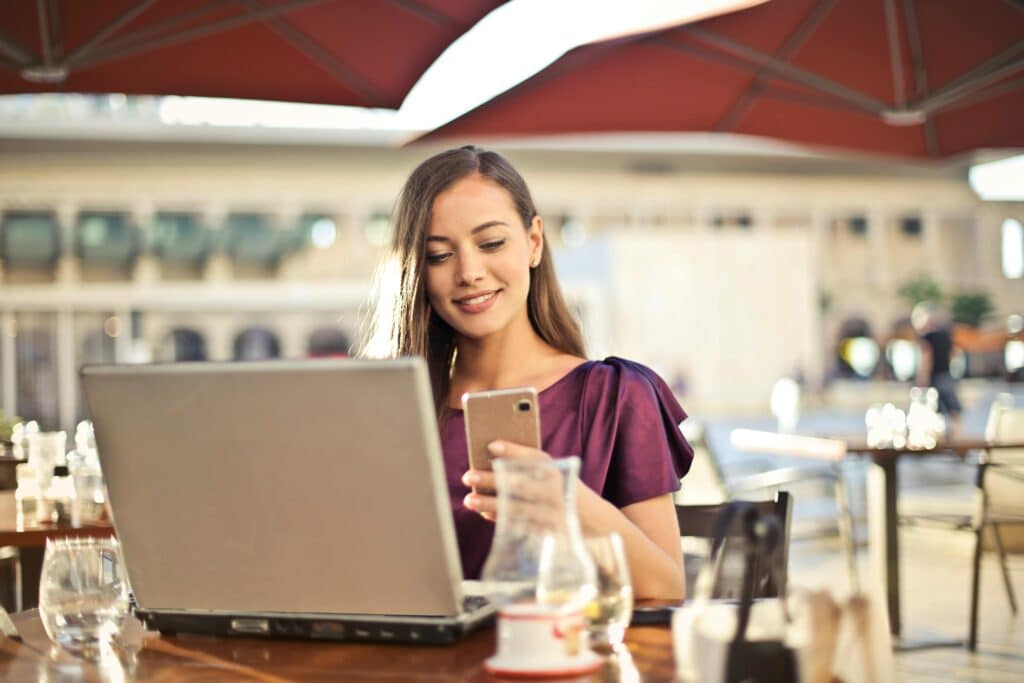 woman-in-purple-shirt-smiling-and-looking-at-phone-while-using-laptop-in-cafe
