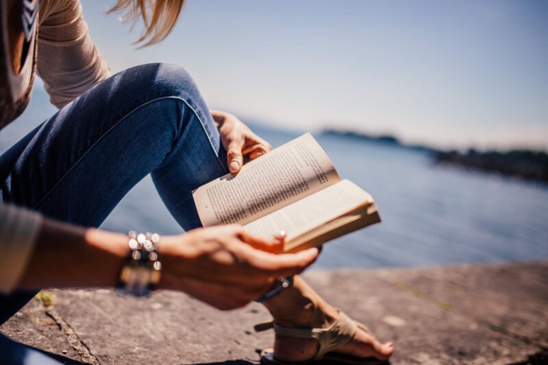 young blonde woman reading a book on the ground next to the ocean