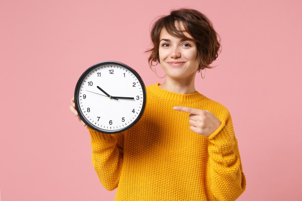 smiling young woman holding a clock and pointing at it