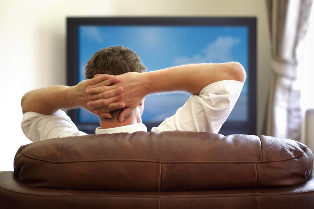 man-resting-on-a-couch-watching-tv-with-his-hands-linked-behind-his-head