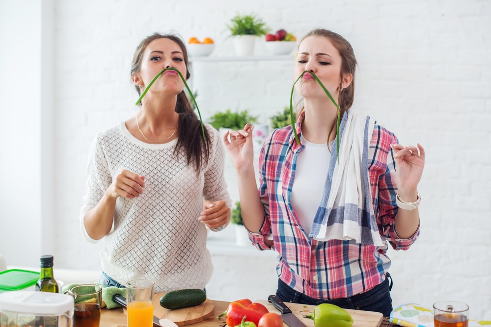two-women-in-a-kitchen-chopping-vegetables-balancing-a-long-green-vegetable-on-their-upper-lips