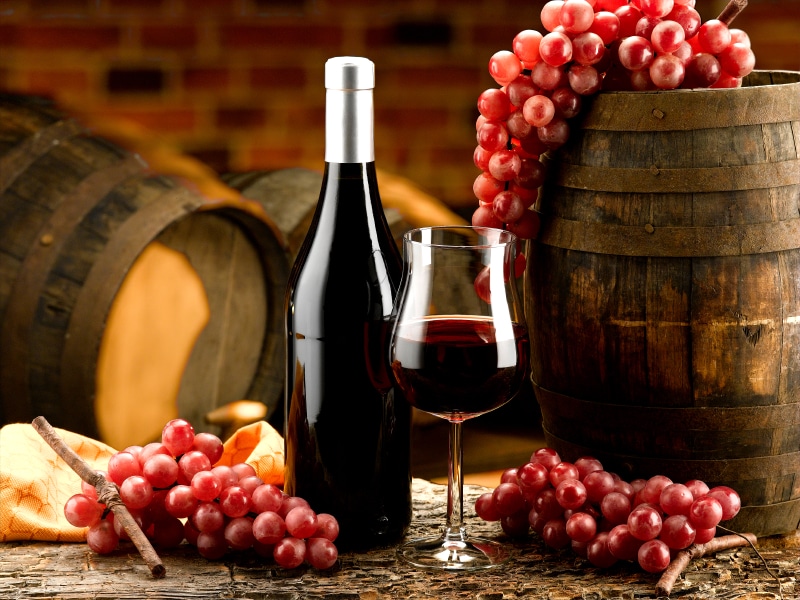 A glass of red wine beside a wine bottle with wine barrels and grapes in the background