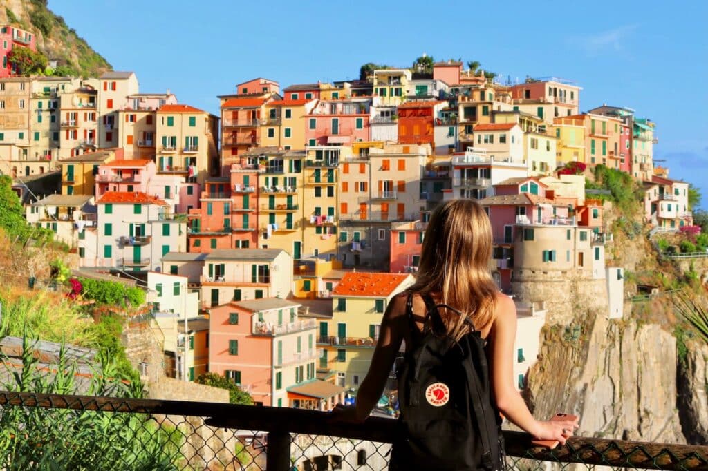 A woman wearing a black backpack, standing at a fence, looking out over the colorful buildings of Manarola, Italy in the distance.