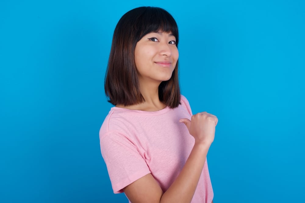 Cheerful Asian woman points at herself with thumb