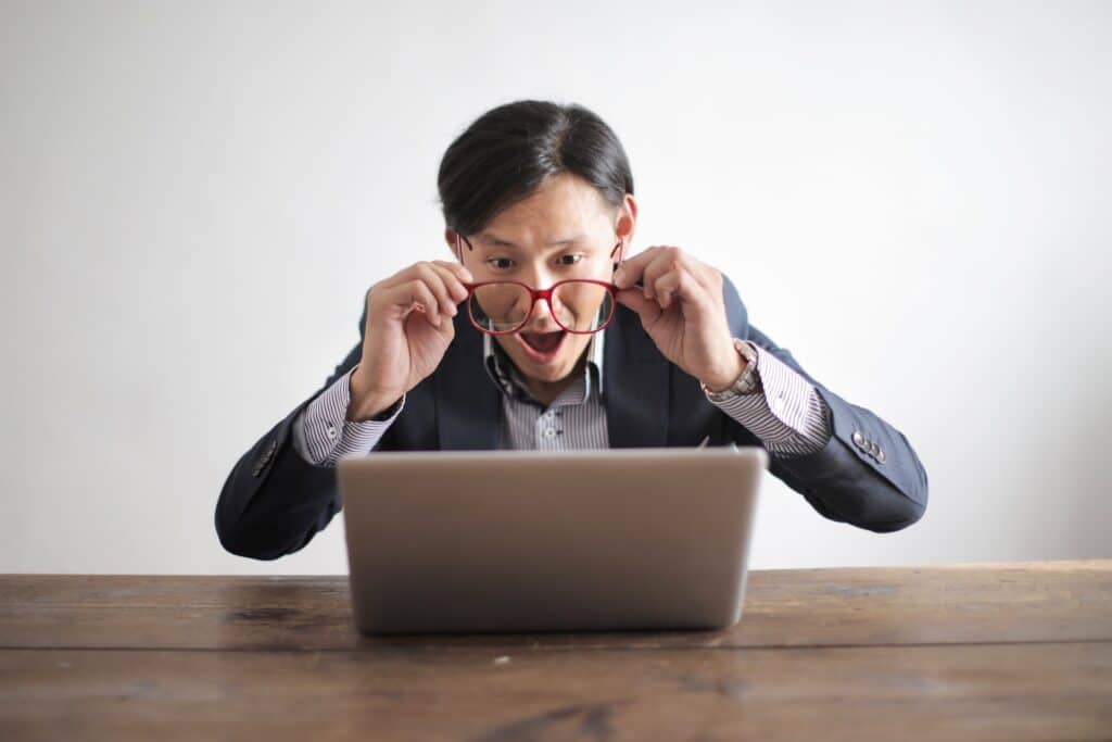 surprised-asian-man-in-formal-wear-taking-off-red-rimmed-glasses-while-looking-at-laptop-screen