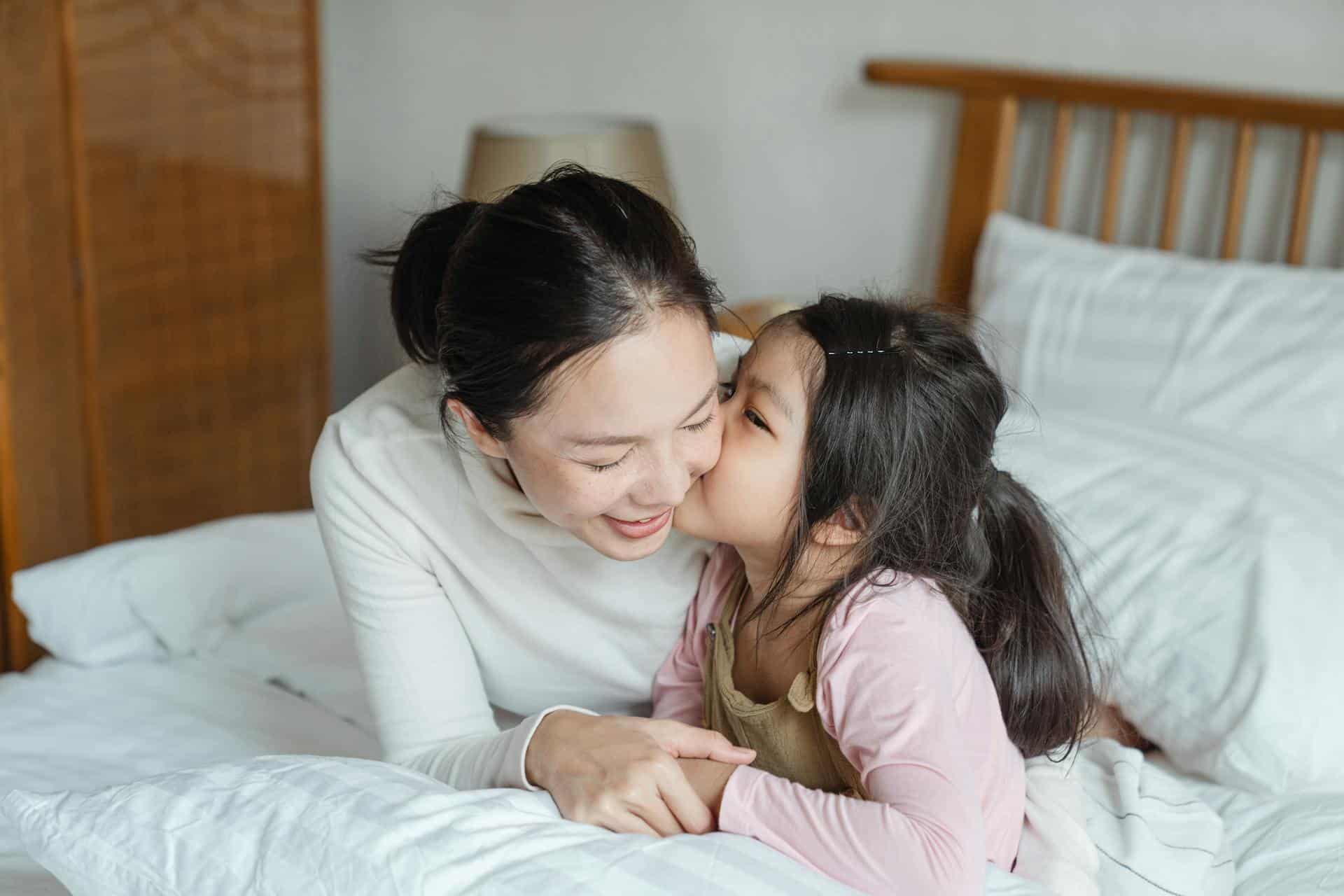 asian-mom-being-kissed-on-the-cheek-by-her-young-daughter-in-bed