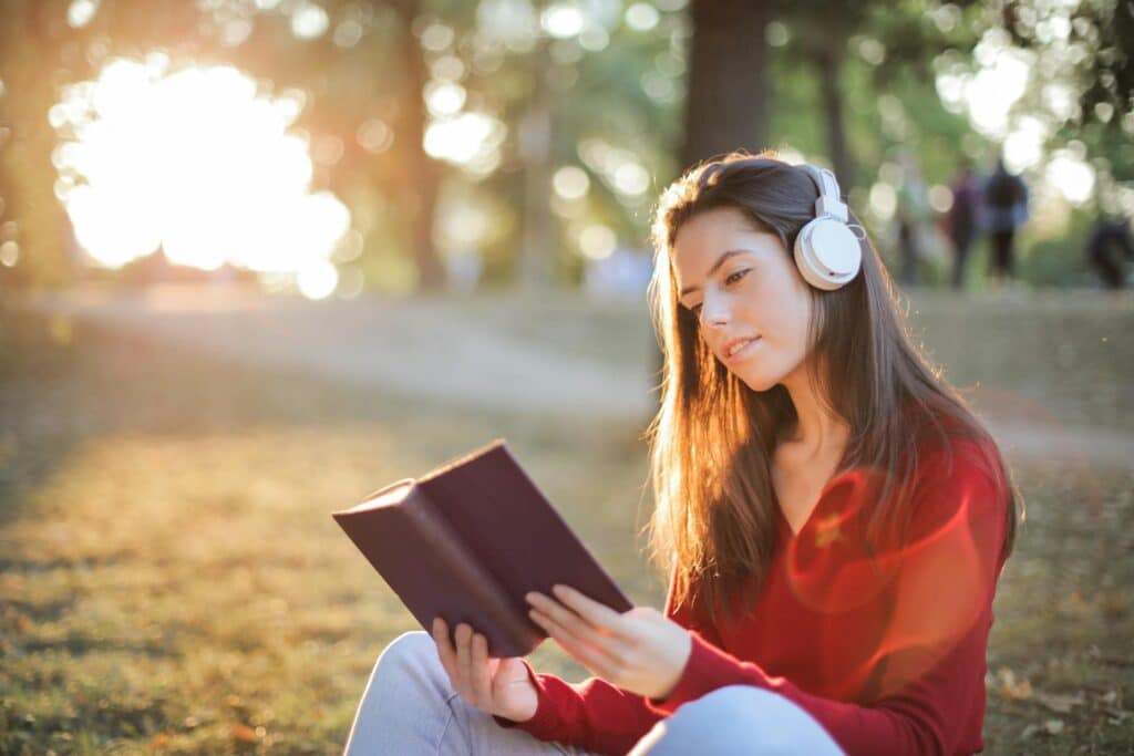 woman-listening-to-white-headset-while-reading-book-near-forest-area