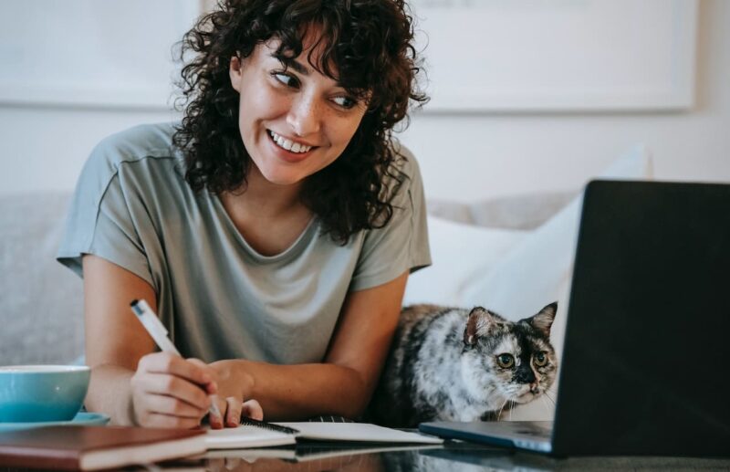 A happy woman (and her marbled-tabby cat) taking notes from a laptop 