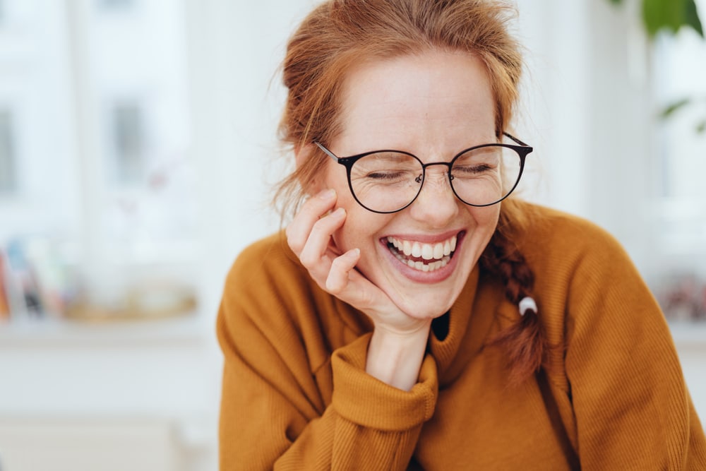Pretty red-haired girl with braid, wearing glasses and orange sweatshirt, laughing with her eyes closed. Close-up front portrait indoors with copy space