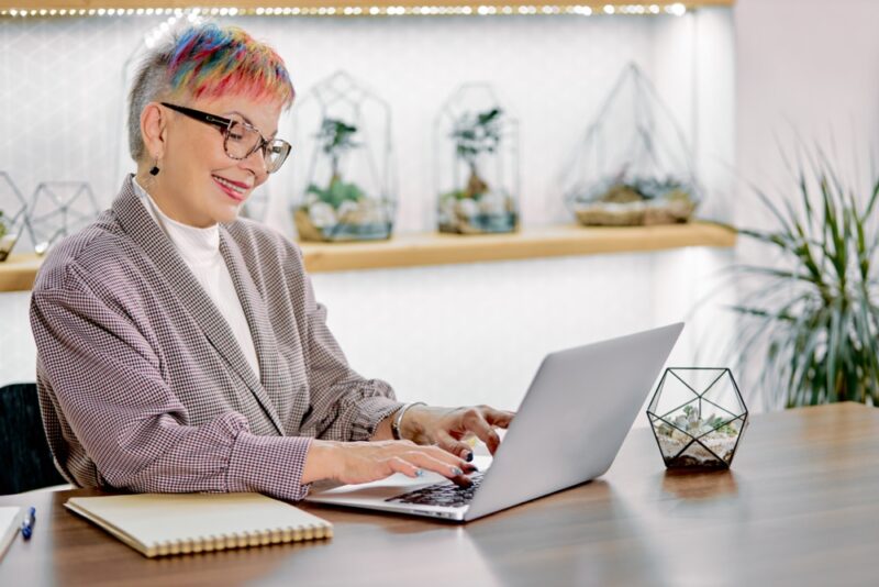 a woman with short colorful hair wearing glasses and blazer sit looking at screen of laptop and typing