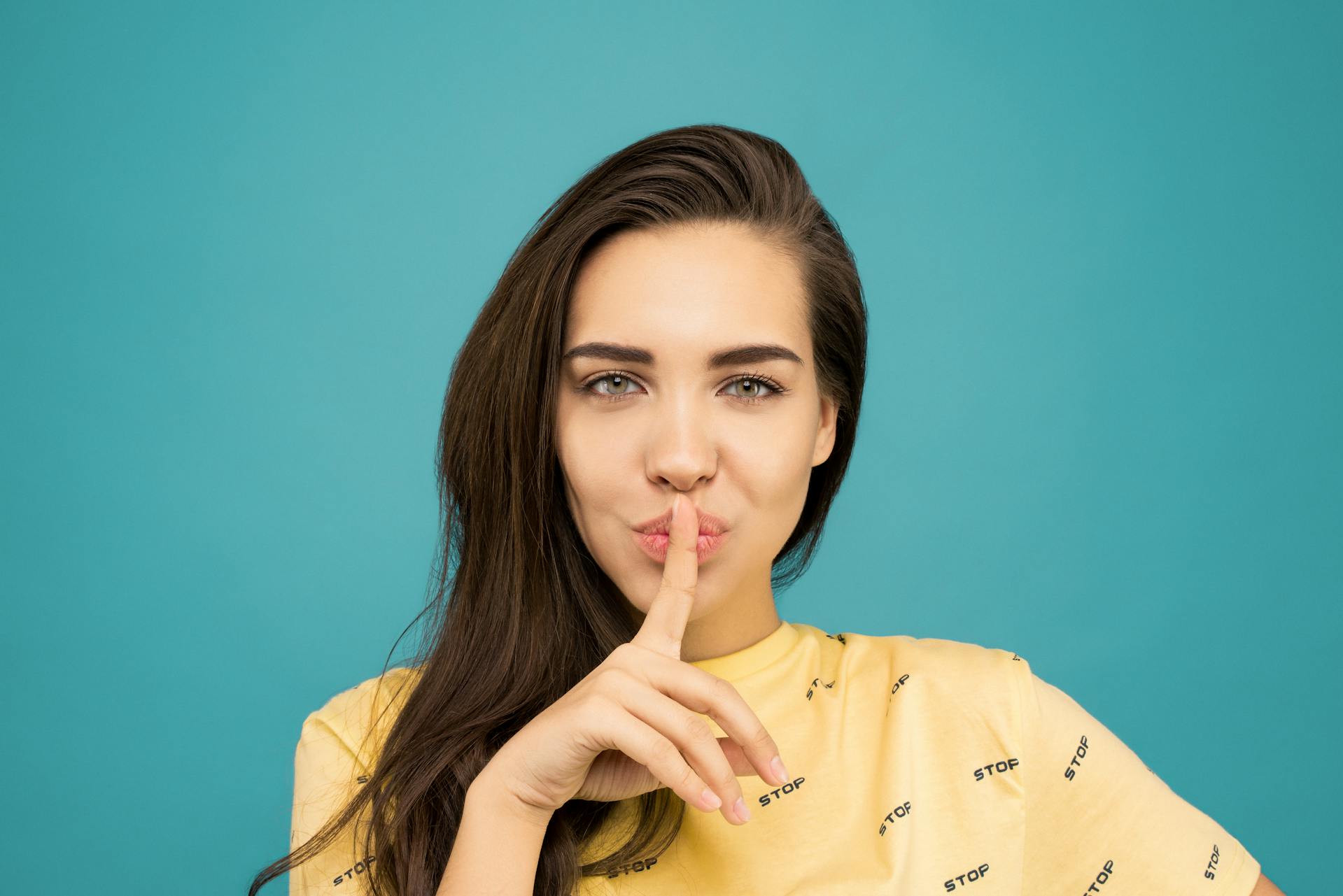 portrait-of-beautiful-woman-in-yellow-shirt-holding-index-finger-to-lips-against-blue-background