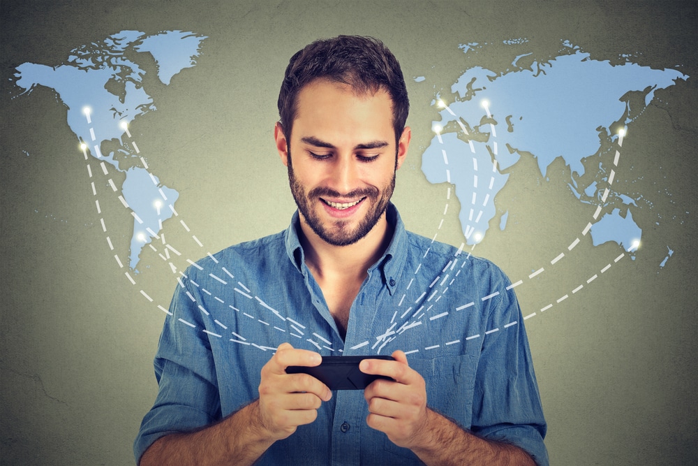 man in blue collared shirt smiling looking at phone with world map connected by dotted lines to the phone in the background