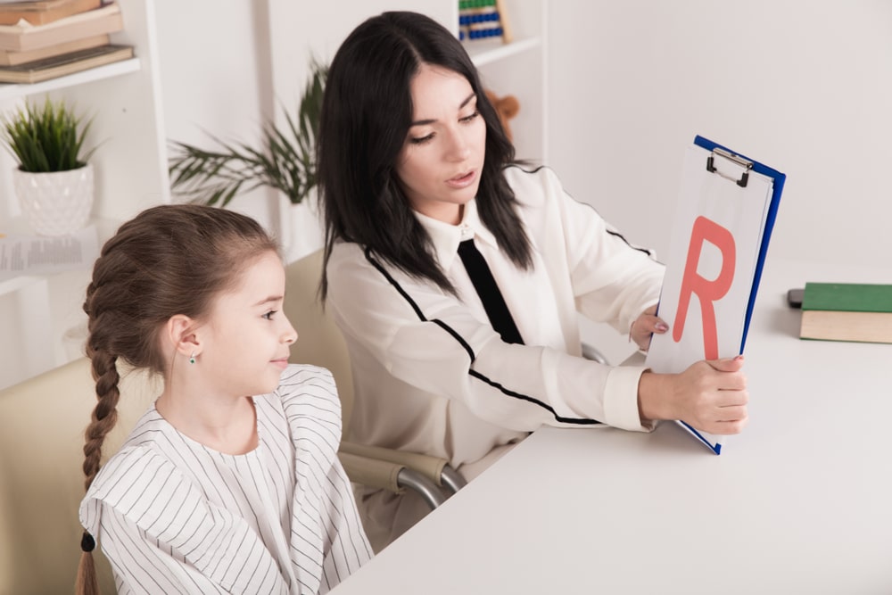 A woman is holding up a large print-out of the letter "R" and practicing pronunciation with a young girl.