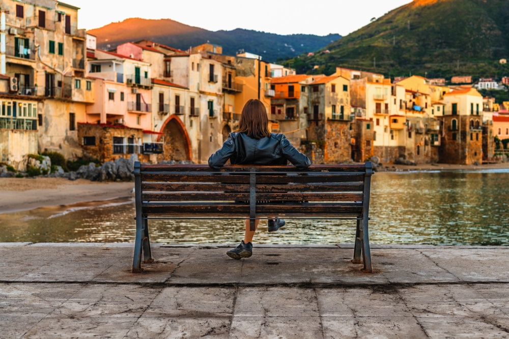 woman sitting on a bench in front of a river looking at the mountains and houses in Italy