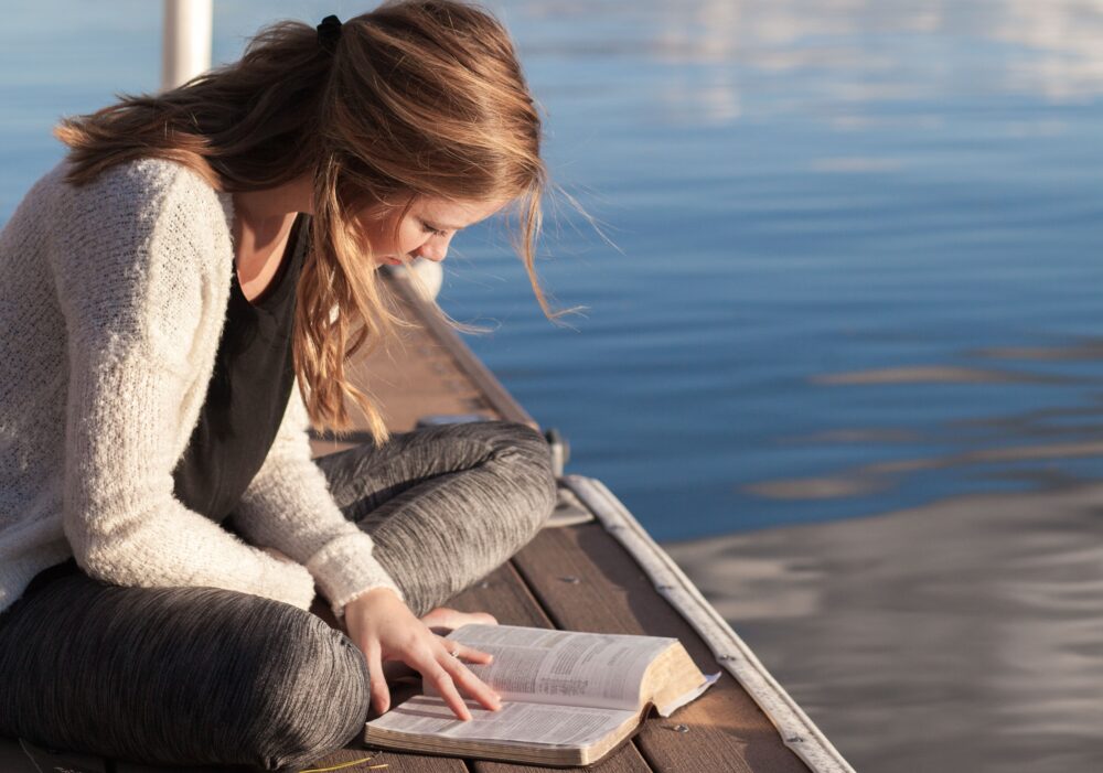 A woman reading a book on a dock by a lake.