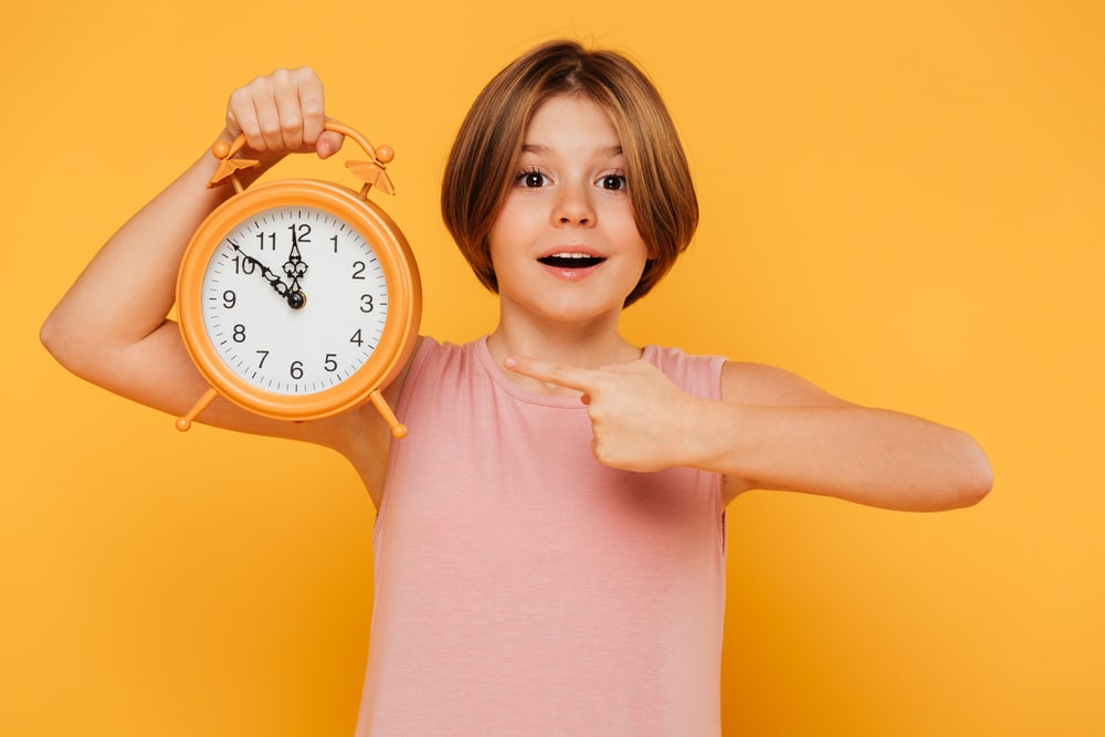 Happy-little-girl-pointing-with-finger-at-alarm-clock-while-looking-at-camera-isolated-over-yellow-background