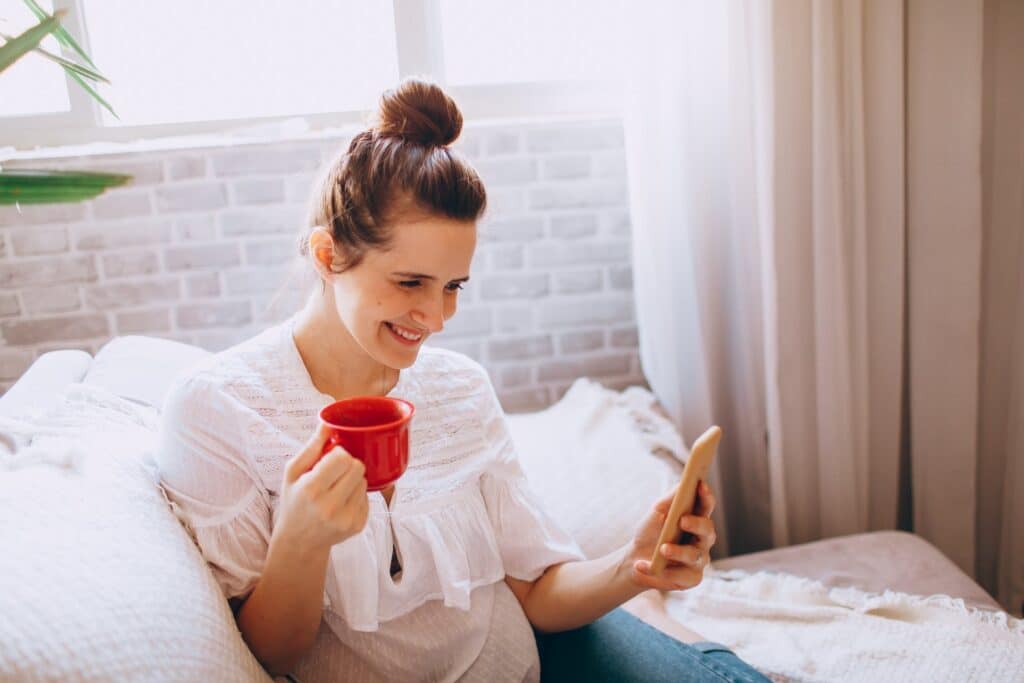 smiling-young-woman-holding-red-cup-in-her-right-hand-and-smartphone-in-her-left-hand